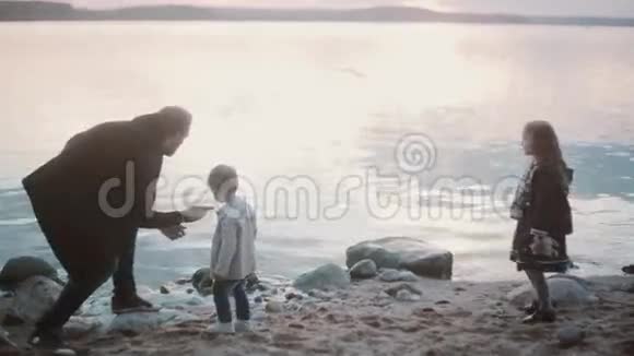 父亲儿子女儿在岸上男人蹲着向男孩和女孩展示如何玩石头跳绳在水上做圆圈视频的预览图