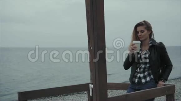 可爱的金发女人穿着黑色皮夹克在海边喝咖啡视频的预览图