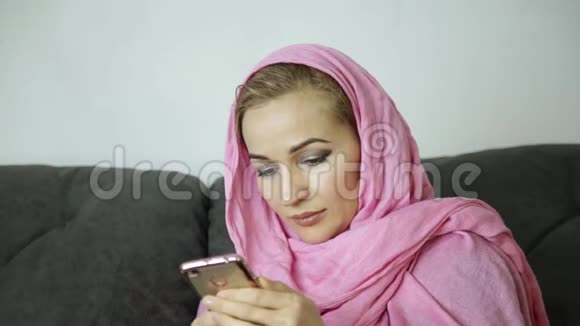 穿着粉红色头巾的年轻穆斯林妇女坐在咖啡馆的沙发上用手机聊天视频的预览图
