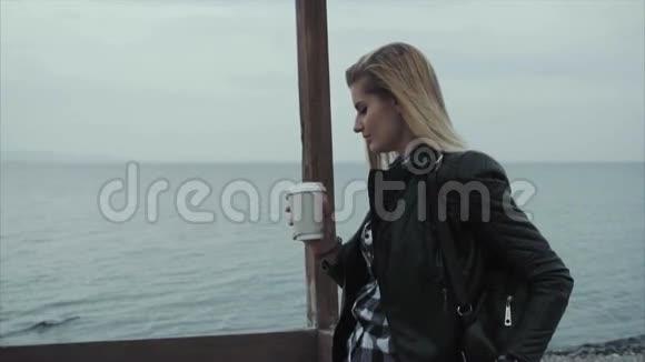 漂亮的金发女人穿着黑色皮夹克在海边喝咖啡视频的预览图