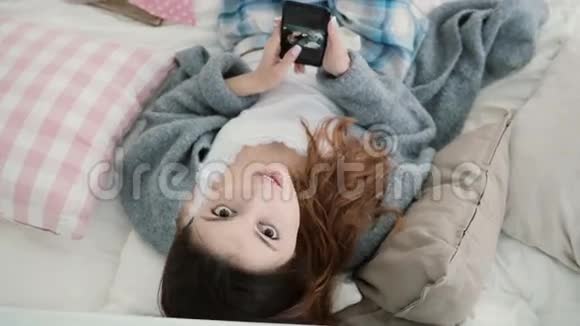 年轻漂亮的女人躺在床上在智能手机上看照片女孩看着相机微笑视频的预览图