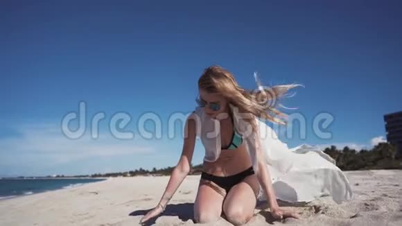 一个穿着彩色比基尼和透明白色斗篷的漂亮女孩坐在沙滩上视频的预览图