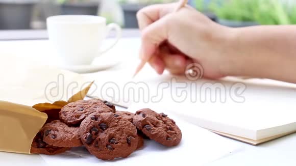 一只手拿着一支木笔写在笔记本上的女性放在一张白色的桌子上抓着吃巧克力饼干视频的预览图