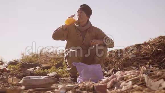 一个肮脏的无家可归的饥饿男人在垃圾堆里喝着包裹里丢失的果汁步行去寻找视频的预览图