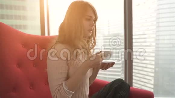 在家中的年轻女性坐在窗前的现代红色椅子上在客厅放松喝咖啡或视频的预览图