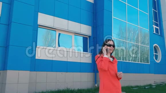 戴墨镜的快乐年轻黑发女人的肖像在旁边的蓝色建筑上打电话视频的预览图