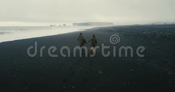 这对年轻夫妇在冰岛黑色火山海滩上奔跑的背面鸟瞰图海边的浪漫约会视频的预览图