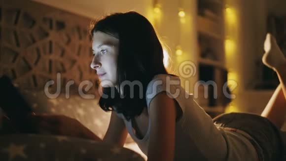 年轻的笑脸女人用平板电脑分享社交媒体晚上躺在家里的床上视频的预览图