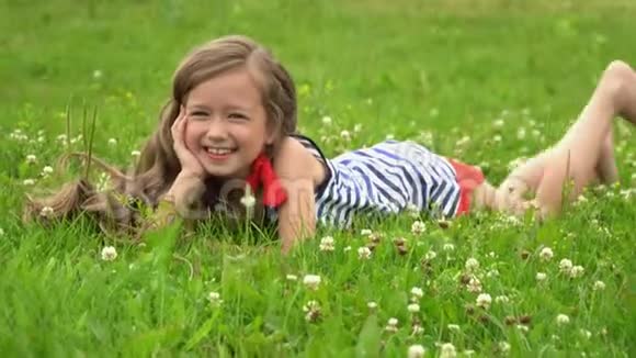 可爱的小女孩坐在绿草上微笑美丽明亮的女孩在相机上摆姿势视频的预览图