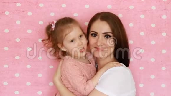 母女拥抱微笑玩得开心笑着亲吻年轻女子和她的孩子在粉色上的特写肖像视频的预览图
