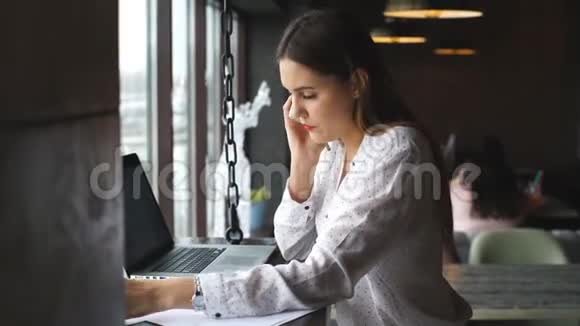 这位年轻的女商人在一家带着笔记本电脑的咖啡馆工作在文件上签名并在电话上交谈视频的预览图
