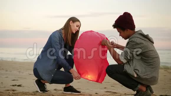 日落时分在海滩上浪漫的约会年轻的多民族夫妇在发射红纸灯笼之前视频的预览图