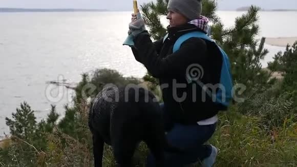一个年轻的女孩走在一条黑狗旁边在靠近水的高坡上在电话里拍了一张美丽的风景照片视频的预览图