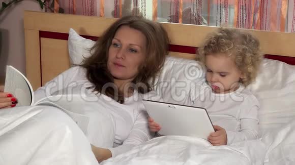 疲惫的母亲在看书她可爱的女儿在平板电脑上看电影视频的预览图