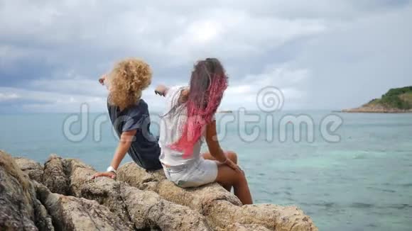 两个年轻的无忧无虑的嬉皮士女孩开始坐在海边的岩石上张开双臂高清慢运动后视图苏视频的预览图