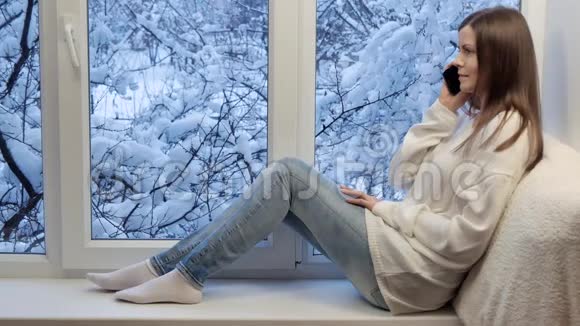 坐在窗台上用智能手机说话的漂亮女孩冬天在外面视频的预览图