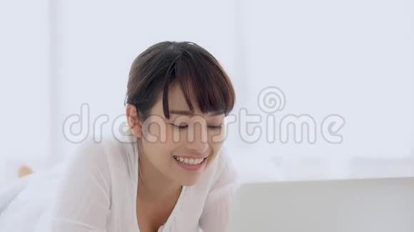 美丽的亚洲年轻女子躺在床上在卧室里用手提电脑上网购物视频的预览图