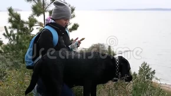 一个戴着裤子的小女孩和一只大狗坐在湖的高岸上手里拿着电话视频的预览图
