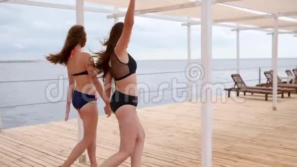 快乐的朋友沿着海边的码头前进长发女孩穿着泳衣手牵着手视频的预览图