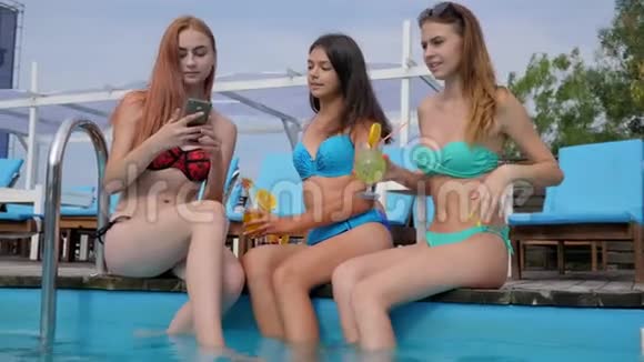 游泳池边穿着带鸡尾酒的泳装女郎自拍年轻女性在小玩意上录制视频视频的预览图