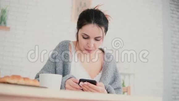 年轻女子在早餐时穿着睡衣使用手机有魅力的女孩在手机上浏览互联网视频的预览图