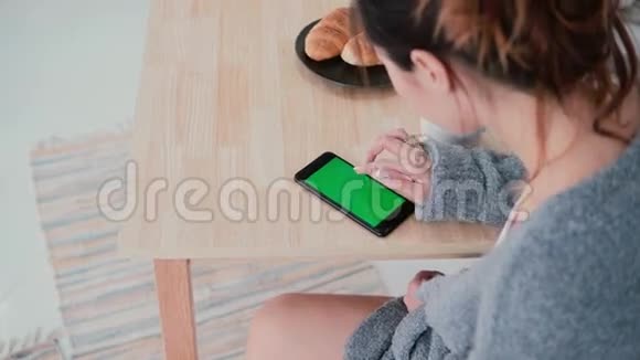年轻女子坐在桌子旁摸着厨房的屏幕女孩早餐时使用智能手机绿色屏幕视频的预览图
