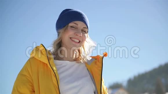 在阳光明媚的冬日里穿着鲜艳的黄色夹克和深色帽子的漂亮金发美女的特写镜头微笑视频的预览图