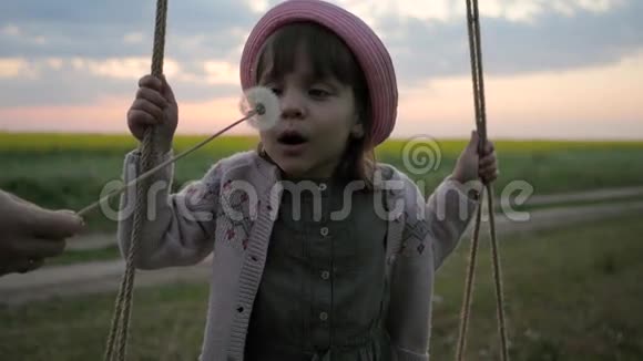 吹蒲公英肖像可爱的女孩骑在秋千上女孩子吹在吹球花快乐的孩子在视频的预览图