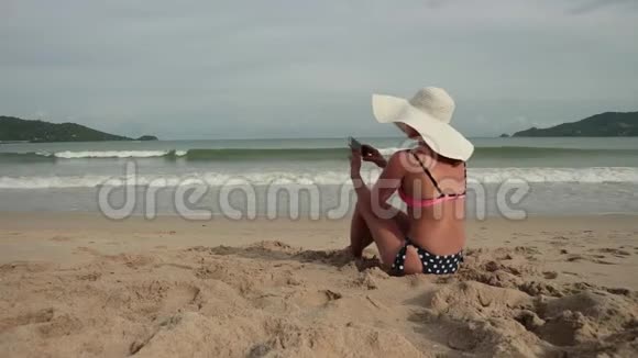 年轻女子在海滩上晒日光浴时用手机穿着比基尼和太阳帽的漂亮女孩拍照视频的预览图