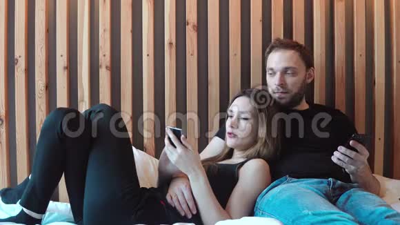 年轻夫妇躺在床上带着智能手机一起休息男人给女人看照片讨论新闻视频的预览图