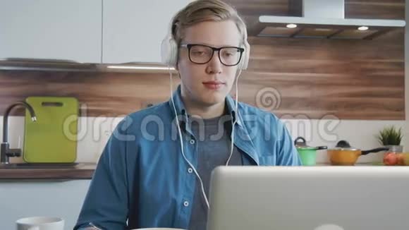 年轻的笑男在家看笔记本电脑上的流媒体电影带着耳机吃麦片视频的预览图