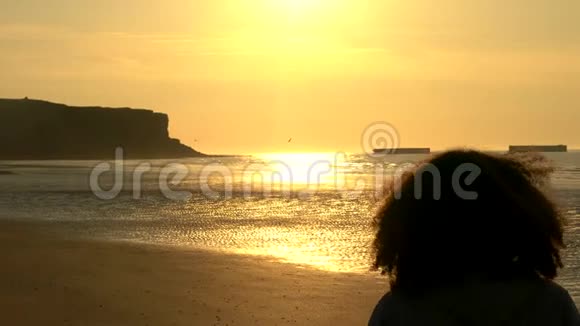 一位留着卷发的年轻女性站在沙滩上看日落或日出视频的预览图