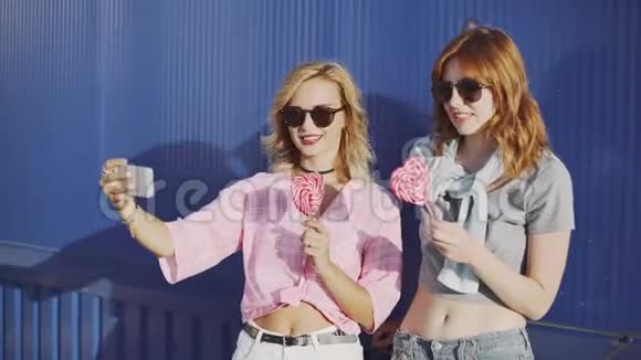 两个年轻漂亮的女人戴着太阳镜聊天吃棒棒糖在街上自拍视频的预览图