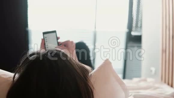 深色头发的女人躺在床上在智能手机上浏览互联网女孩呆在家里视频的预览图