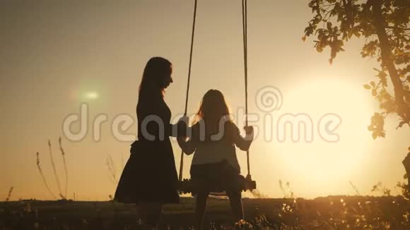 幸福的年轻母亲和小女儿在阳光下荡秋千的剪影坐在木秋千上的漂亮女孩视频的预览图