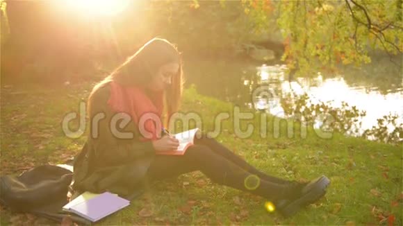 美丽的女孩坐在草地上写着一本笔记本背景是美丽的公园和湖视频的预览图