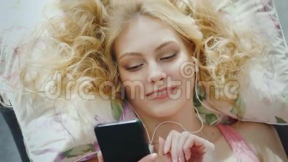 一个迷人的年轻女人躺在床上的肖像使用智能手机听音乐从上面看视频的预览图