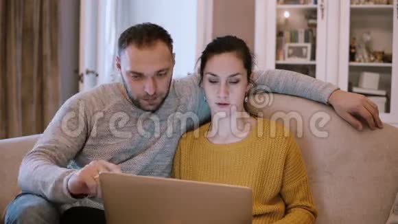 年轻夫妇坐在沙发上拿着电脑男人和女人坐在客厅里使用笔记本电脑视频的预览图