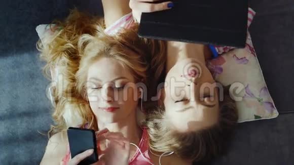 两个年轻的女人在家里从设备上放松听音乐使用手机和平板电脑坐在头旁边视频的预览图