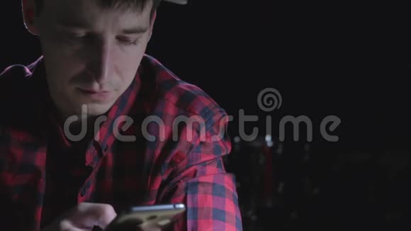 穿着红色格子衬衫的疲惫和压力很大的年轻人厌倦了在黑暗的背景下用手机上网视频的预览图