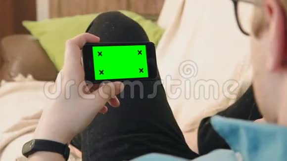 躺在沙发上看着绿色屏幕的智能手机的年轻人视频的预览图