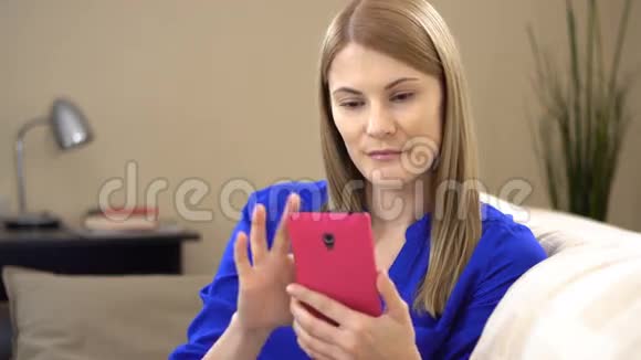 漂亮的年轻女人穿着蓝色t恤智能手机坐在沙发上浏览互联网微笑视频的预览图
