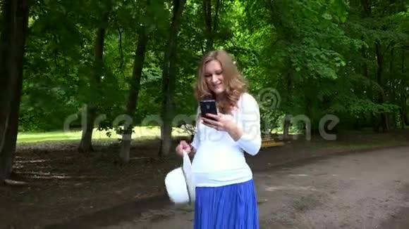 微笑可爱的孕妇手里拿着帽子和电话走在公园里视频的预览图