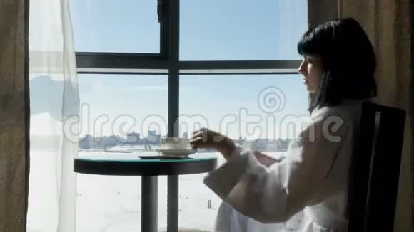 年轻漂亮的女人坐在靠窗的桌子旁喝茶喝咖啡用手机发信息视频的预览图