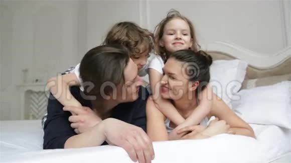 快乐的父母和两个爱笑的孩子正躺在白色的床上在周末的家里一起玩帅哥视频的预览图