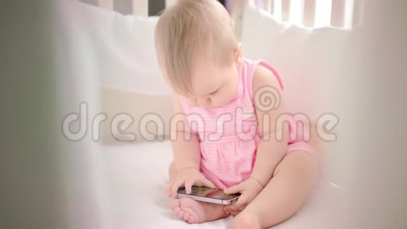 婴儿床上有电话的小女孩幼儿在手机上观看动画片视频的预览图