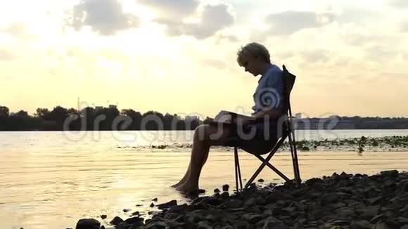年轻人坐在河岸上写作把脚留在水里视频的预览图