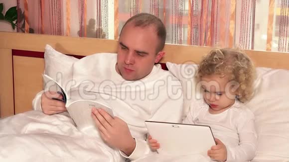 疲惫的父亲读着书可爱的女儿在平板电脑上看电影视频的预览图