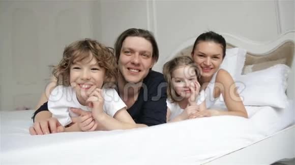 有两个孩子的年轻微笑的父母在他们的卧室里一起玩欢乐的家庭在白色的床上视频的预览图