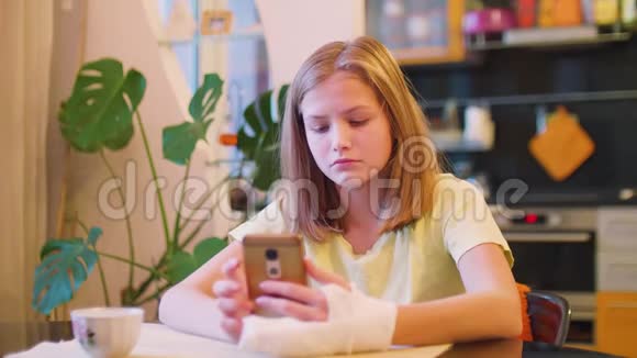 年轻的白种人金发碧眼左臂骨折端着智能手机坐在桌子旁视频的预览图